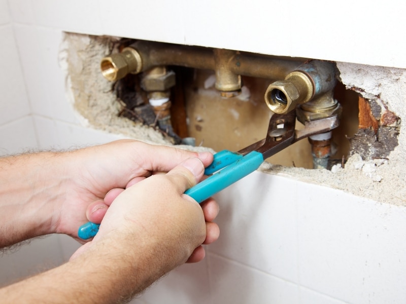Close-up of hands using pliers for DIY shower leak repairs on exposed pipes in a tiled wall.